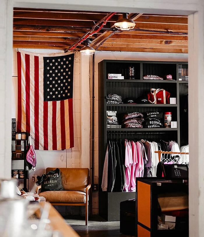 Inside Roswell Supply Co. at The Webb: an American flag, a leather armchair, shelves of Georgia caps and sweatshirts and a clothing rack under a wood-beamed ceiling